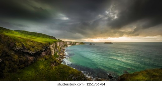 Scenic View At The Steep Coastline Of Ballintoy/Carrick-a-Rede In Ireland, With A Dark Clouded Sky Above A Green Sea Seen From The Side Of The Famous Rope Bridge