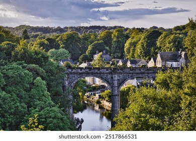 A scenic view of a stone bridge arching over a river, surrounded by lush greenery and quaint houses. In Knaresborough - Powered by Shutterstock