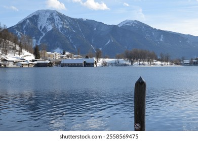 Scenic view of Tegernsee Lake with snow-covered mountains in the background, reflecting on calm waters. Wooden pier and "no swimming" sign in the foreground - Powered by Shutterstock