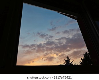 Scenic view through an open window frame showing a colorful sunset sky filled with soft pastel clouds and silhouettes of tree tops. Concept of staycation, hope, tranquil evening, and home view.