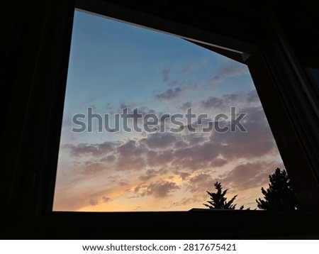 Scenic view through an open window frame showing a colorful sunset sky filled with soft pastel clouds and silhouettes of tree tops. Concept of staycation, hope, tranquil evening, and home view.