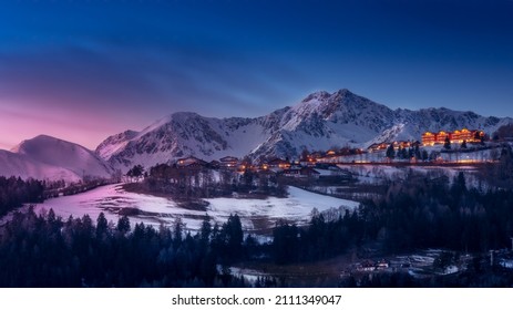 Scenic view of a town with hotels against blue sky and snowcapped mountains at sunset in winter. Snow and forest in the foreground - Shutterstock ID 2111349047
