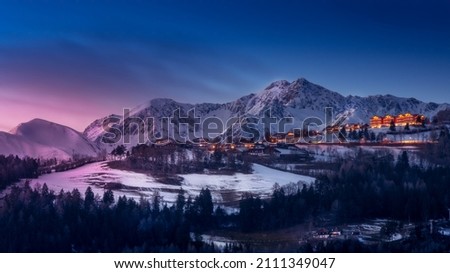 Scenic view of a town with hotels against blue sky and snowcapped mountains at sunset in winter. Snow and forest in the foreground