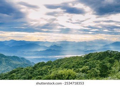 A scenic view of a valley with lush greenery in the foreground and multiple mountain ranges in the distance under a cloudy and sunlight sky.  - Powered by Shutterstock