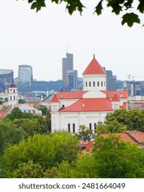 A scenic view of Vilnius, Lithuania, featuring a prominent historic church with a red roof amidst lush greenery and modern skyscrapers in the background