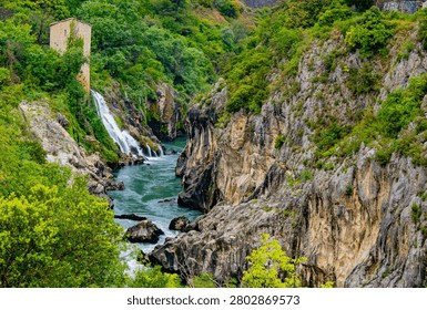 Scenic view of a waterfall cascading down rocky cliffs surrounded by lush greenery, with a historical tower nearby, creating a picturesque landscape.