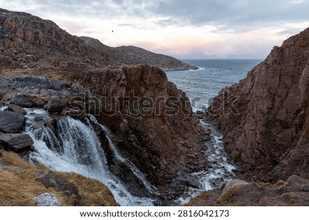 Scenic view of a waterfall cascading down red rocky cliffs toward the ocean bay in northern arctic landscape during twilight.