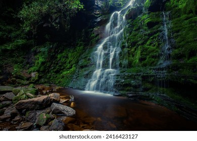 A scenic view of waterfall to the rocky river under green mountains in the forest within trees - Powered by Shutterstock