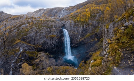 A scenic waterfall cascades down a rocky cliff surrounded by autumn foliage in a mountainous landscape. - Powered by Shutterstock