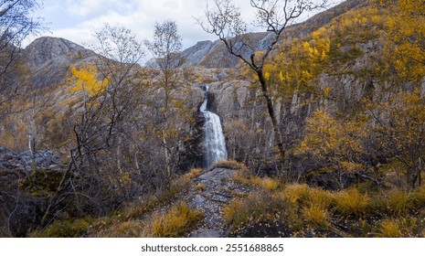 A scenic waterfall cascades down a rocky cliff surrounded by autumn foliage and barren trees in a mountainous landscape. - Powered by Shutterstock