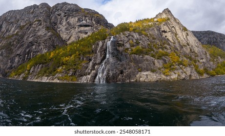 A scenic waterfall cascading down a rocky cliff into a calm body of water, surrounded by rugged mountains and autumn foliage under a cloudy sky. - Powered by Shutterstock