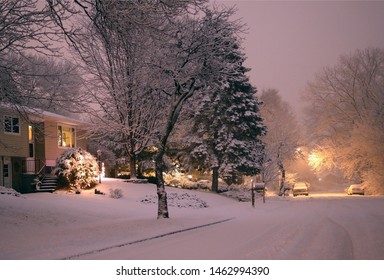 Scenic Winter Blizzard Evening Landscape. Small Town Street View During Snowfall At Dusk With Parked Cars And Houses With Decorated For Christmas Front Yards.