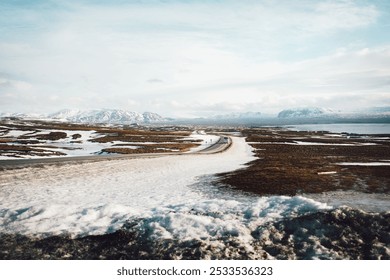 A scenic winter landscape featuring a winding road through snow-covered fields and distant mountains under a partly cloudy sky. - Powered by Shutterstock