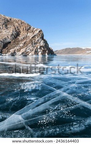 Scenic winter landscape of frozen Baikal Lake with blue transparent ice with cracks and bubbles near Shibete Rock and bay of Anga River on sunny February day. Winter travel on ice. Natural background