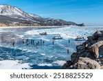 A scenic winter landscape of people walking and exploring a frozen lake surrounded by snow-covered mountains. Hovercrafts and groups of adventurers stand on the transparent ice. Lake Baikal in winter.