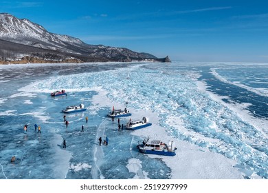 A scenic winter landscape of people walking and exploring a frozen lake surrounded by snow-covered mountains. Hovercrafts and groups of adventurers stand on the transparent ice. Lake Baikal in winter.