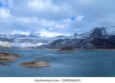 Scenic winter landscape of a snow-covered mountain range and calm lake, with dramatic clouds overhead, creating a serene atmosphere. - Powered by Shutterstock