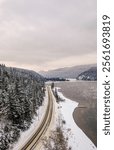 A scenic winter road winds along a snowy lakeshore surrounded by forested hills under a overcast sky, depicting serene and tranquil nature in British Columbia.