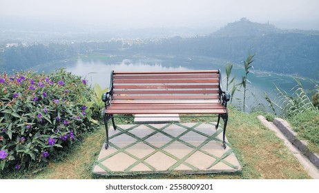 A scenic wooden bench with wrought-iron details placed on a lush green hilltop, overlooking a serene lake and distant hills shrouded in mist. - Powered by Shutterstock