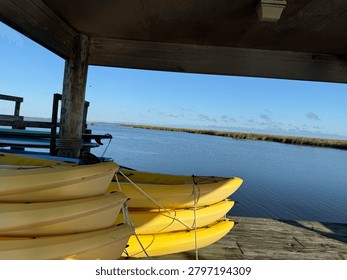 A scenis view from under a rustic, covered wooden dock showing stacks of yellow rental kayaks parked on the shoreline. It is next to a calm blue inlet under a brigh sky.