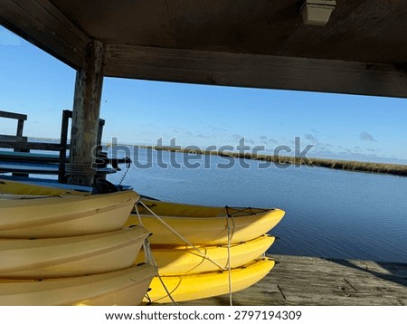 A scenis view from under a rustic, covered wooden dock showing stacks of yellow rental kayaks parked on the shoreline. It is next to a calm blue inlet under a brigh sky.