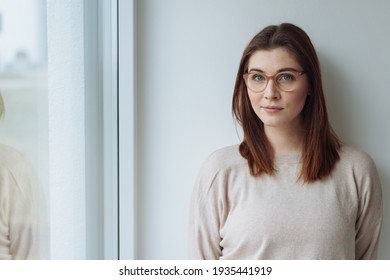 Sceptical young woman staring thoughtfully at the camera with a serious dubious expression as she stands alongside a window indoors with copyspace - Shutterstock ID 1935441919