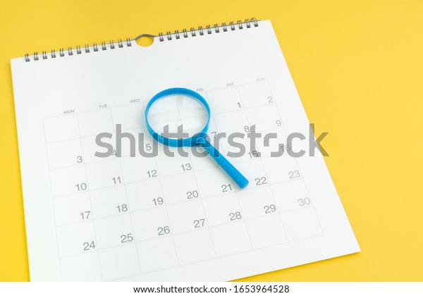 Schedule, focus date or special event day\
concept, blue magnifying glass on white clean calendar on solid\
yellow background.