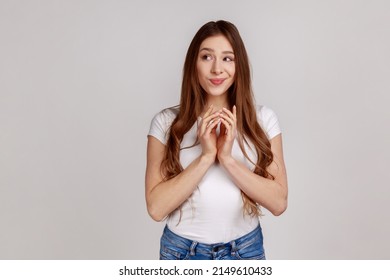 Scheming devious attractive woman clasping hands and thinking over tricky plan, having sly cunning idea to prank, wearing white T-shirt. Indoor studio shot isolated on gray background.