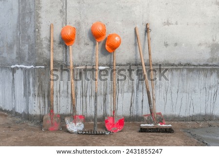 Schippen, Rechen und ein Besen mit Bauarbeterhelmen vor einer Betonwand