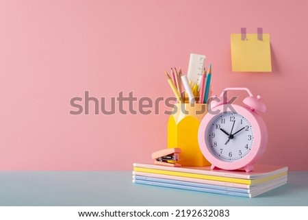 School accessories concept. Photo of stationery on blue desk stand for pens pencils alarm clock stack of notebooks mini stapler and sticky note paper attached to pink wall with empty space