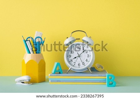 School accessories concept. Photo of stationery on blue desk stand for pencils alarm clock plastic alphabet letters stack of notebooks stapler and adhesive tape on yellow wall background