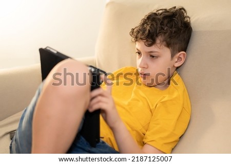 School aged kid using a tablet relaxing on the coach indoor. Children addicted from technology studying online from home - education, addiction, people and tech concept