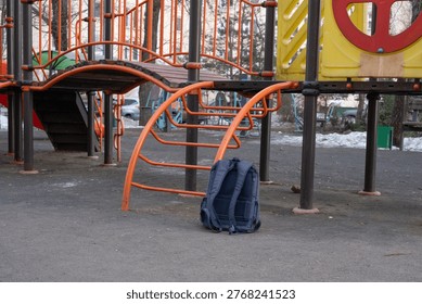School backpack left on the ground beneath a playground structure, symbolizing a child's temporary departure for active play after lessons and the everyday reality of misplaced belongings