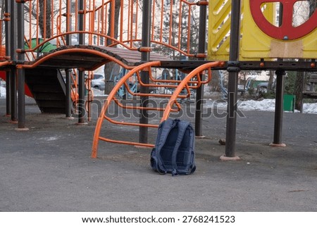 School backpack left on the ground beneath a playground structure, symbolizing a child's temporary departure for active play after lessons and the everyday reality of misplaced belongings
