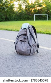 School Backpack With Medical Mask On The Ground, Green Soccer Field And Sunset Background. Back To School During Epidemic Concept. 