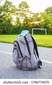 School Backpack With Medical Mask On The Ground, Green Soccer Field And Sunset Background. Back To School During Epidemic Concept. 