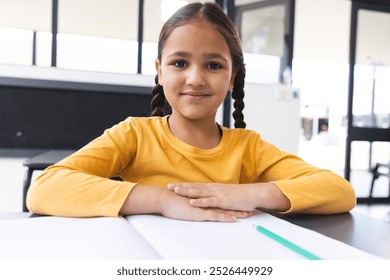 In school, biracial young girl sitting at a desk in a classroom, smiling at camera. She has dark braided hair, light brown skin, and is wearing a yellow top, unaltered - Powered by Shutterstock