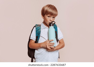 School boy with backpack drinks water bottle at studio isolated over beige background. Childhood, five years child, kid, excursion, education concept. Back to school. - Powered by Shutterstock