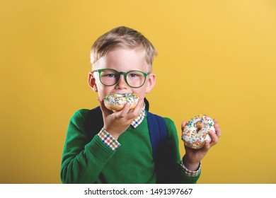 School Boy Is Eating Donut On Yellow Background Wall. Child Has Snack With Junk Food. Stylish Kid At Class With Bag. Back To School