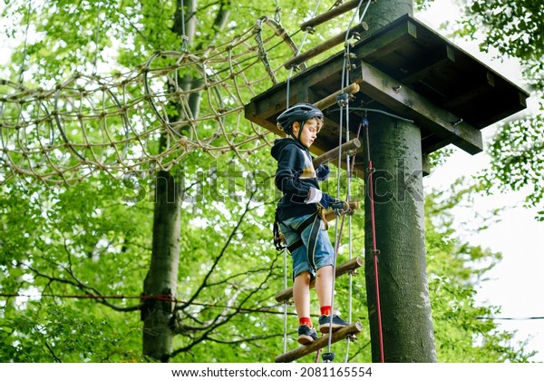 School boy in forest adventure park. Acitve\
child, kid in helmet climbs on high rope trail. Agility skills and\
climbing outdoor amusement center for children. Outdoors activity\
for kid and families.