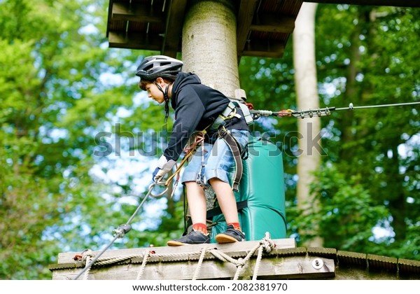 School boy in forest adventure park. Acitve\
child, kid in helmet climbs on high rope trail. Agility skills and\
climbing outdoor amusement center for children. Outdoors activity\
for kid and families.