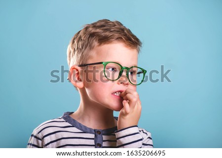 School boy nervously biting nails. Bulling and childhood psychology concept, Child boy in uniform, glasses and backpack isolated on blue. Kid portrait with finger in mouth