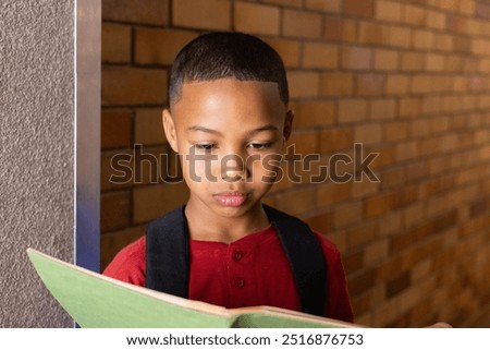 In school, boy reading notebook while standing against brick wall. Education, learning, studying, childhood, literature, student