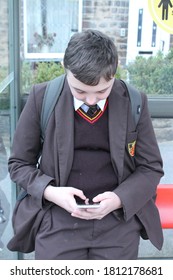 School Boy In School Uniform Sat Texting On A Cell Phone At A Bus Stop.  Lancashire, UK, 09-10-2020