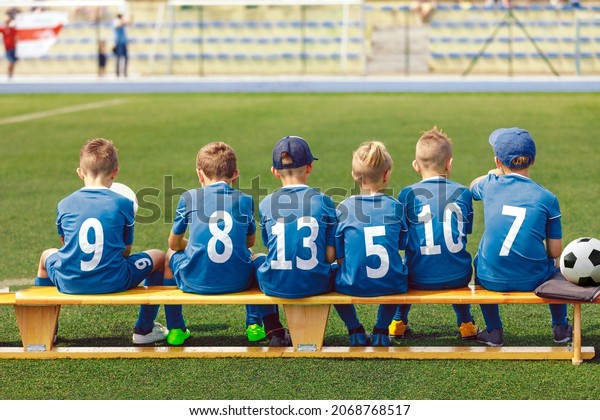 School Boys\
in Sports Football Team. Kids is Classic Blue Soccer Jersey\
Uniforms With Numbers. Kids Playing Sports Together. Children\
Sitting on Sideline Wooden Substitiute\
Bench