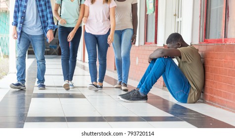 School bullying. Afro american male teenager desperate seated in school hallway while group of teenagers walk towards him.