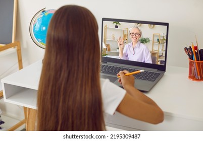 School Child Having Online Class. Little Girl Sitting At Table, Looking At Screen Of Laptop Computer, Watching Video Lecture, Or Having Distant Lesson With Her Teacher. View Over The Shoulder