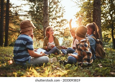 School Children Learning About Ecology, Nature And Environment With Teacher While Sitting Together On Green Grass In Forest On Sunny Day, Kids Raising Hands Asking Questions During Outdoor Lesson