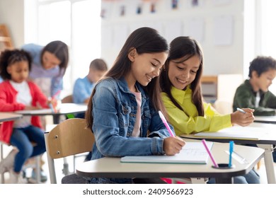 School children sit at desks, writing in copybooks, one girl glances at her friend's test and smiling, studying at school - Powered by Shutterstock