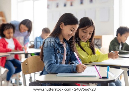 School children sit at desks, writing in copybooks, one girl glances at her friend's test and smiling, studying at school